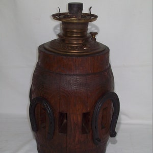 Puede incluir: Una lámpara de aceite de madera con un quemador de latón y un diseño decorativo. La lámpara tiene forma de barril y tiene dos herraduras a los lados.