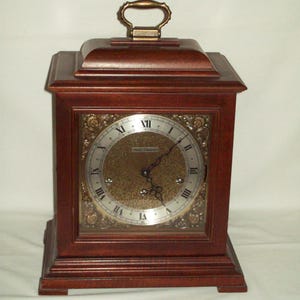 May include: A wooden mantel clock with a rectangular shape and a brass handle. The clock face is gold with black Roman numerals and ornate detailing. The clock is made of dark brown wood.