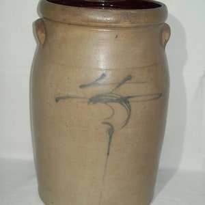 May include: A large, brown stoneware crock with two handles and a blue and black abstract design. The crock is a traditional style of pottery.