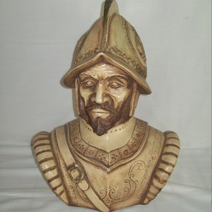 May include: A ceramic bust of a man wearing a helmet and armor. The bust is painted in shades of brown and beige.