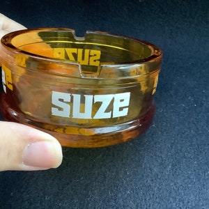 May include: Amber glass ashtray with the word "SUZE" printed in white on the side.