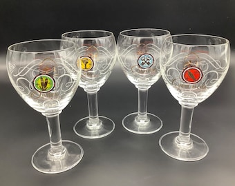 May include: Four clear stemmed wine glasses with decorative swirls and colorful circular logos. Each glass has a different logo with text. The glasses are on a dark surface.