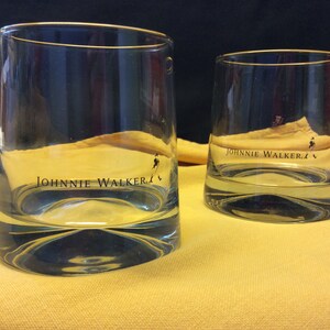 May include: Two clear glass tumblers with a gold rim and the words "JOHNNIE WALKER" printed on the front. The glasses are sitting on a yellow surface.