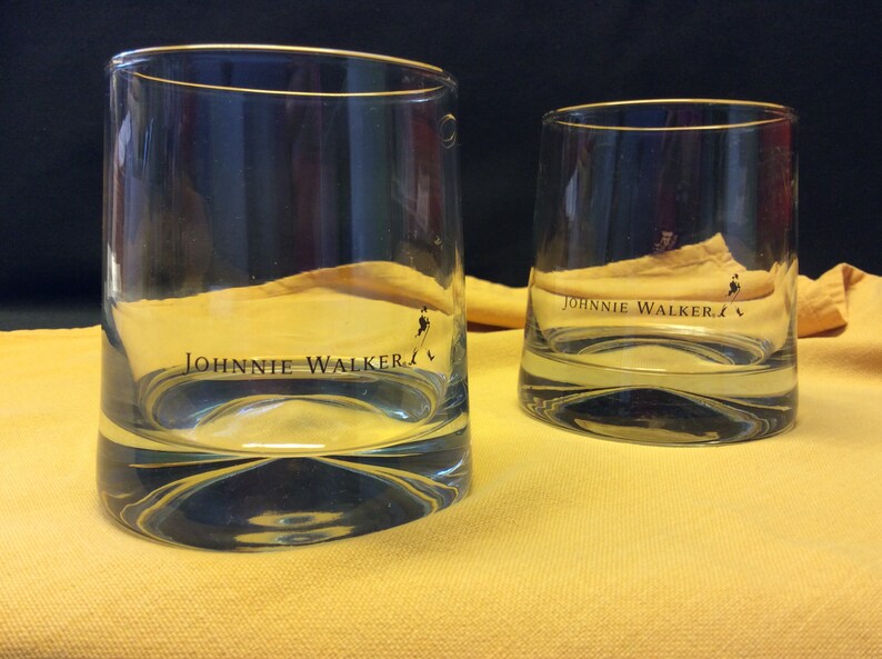 May include: Two clear glass tumblers with a gold rim and the words "JOHNNIE WALKER" printed on the front. The glasses are sitting on a yellow surface.