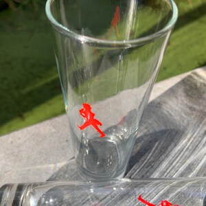 Op de afbeelding: Twee heldere glazen drinkglazen met een rood lopend figuurlogo op elk. De glazen staan op een houten oppervlak.
