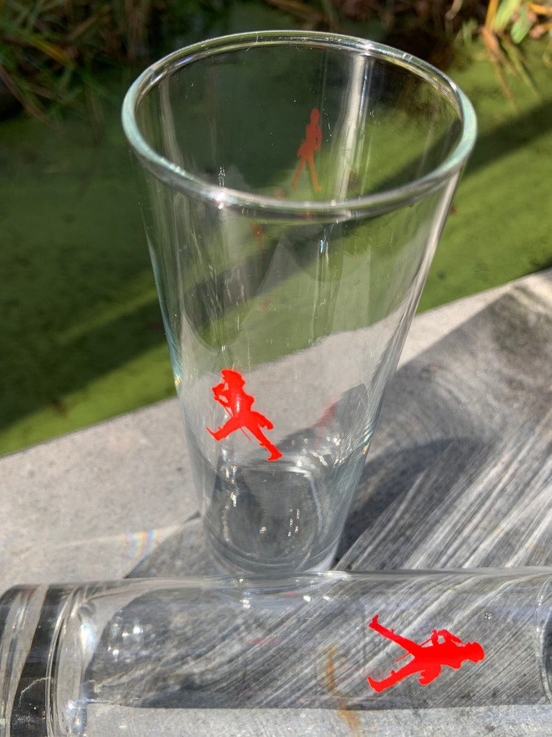 Op de afbeelding: Twee heldere glazen drinkglazen met een rood lopend figuurlogo op elk. De glazen staan op een houten oppervlak.