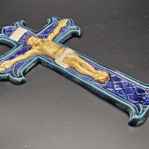 Op de afbeelding: Een keramisch kruisbeeld met een blauw en wit kruis en een beige figuur van Jezus. Het kruis heeft een ruitpatroon en decoratieve randen. De figuur staat in een traditionele pose.