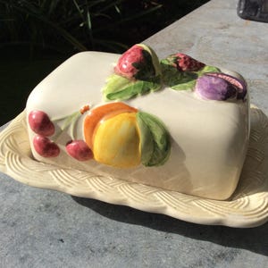 Puede incluir: Un mantequillero de cerámica blanca con tapa decorada con fruta pintada, incluyendo fresas, cerezas, un melocotón y una pera. El plato tiene un patrón tejido alrededor del borde.