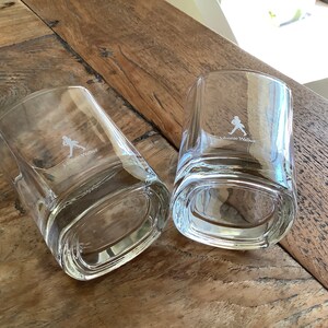 May include: Two clear glass tumblers with a square base and rounded edges. The glasses are etched with the Johnnie Walker logo and a walking man silhouette.