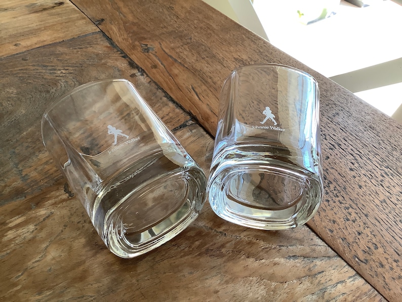 May include: Two clear glass tumblers with a square base and rounded edges. The glasses are etched with the Johnnie Walker logo and a walking man silhouette.