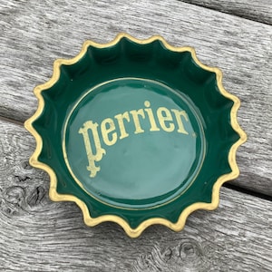May include: A green ceramic dish in the shape of a bottle cap, with a gold rim and the word "perrier" in gold lettering. The dish is on a weathered wooden surface.