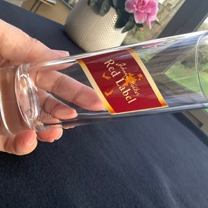 May include: A clear glass shot glass with a red and gold label that reads "Johnnie Walker Red Label".