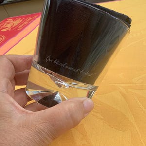May include: A clear, faceted glass with the inscription "Our blend cannot be beat." The glass is partially filled with a clear liquid and topped with a dark, folded cloth. The glass is held against a yellow background.
