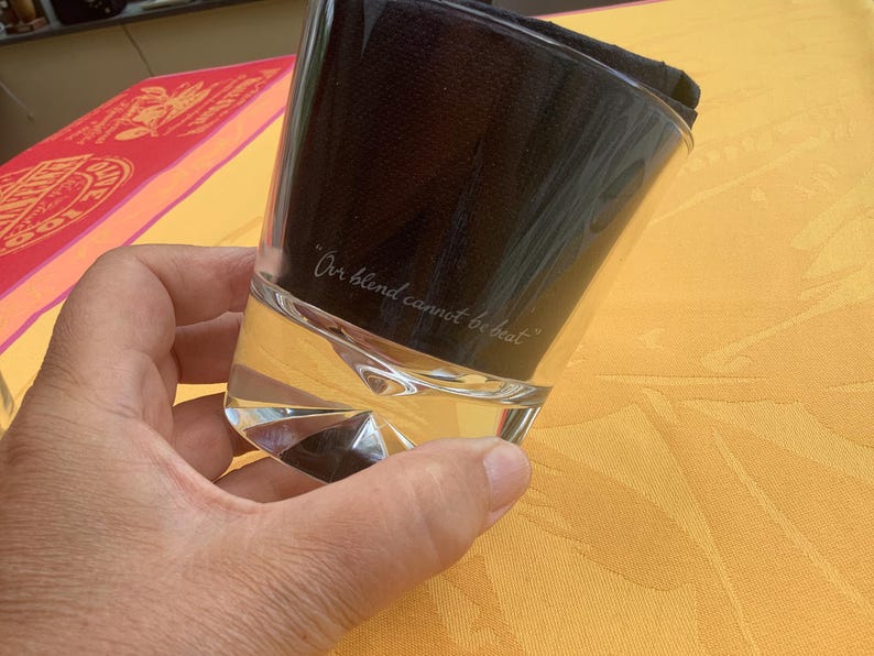 May include: A clear, faceted glass with the inscription "Our blend cannot be beat." The glass is partially filled with a clear liquid and topped with a dark, folded cloth. The glass is held against a yellow background.
