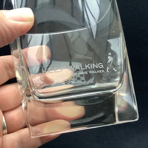 May include: A clear glass tumbler with the words "Keep Walking Johnnie Walker" etched on the side. The glass is empty and has a square shape.
