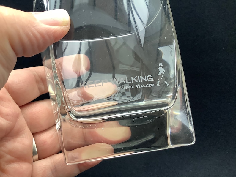 May include: A clear glass tumbler with the words "Keep Walking Johnnie Walker" etched on the side. The glass is empty and has a square shape.