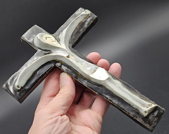 May include: A ceramic cross sculpture with a figure of Jesus. The cross is dark brown with white and light gray accents. The figure is white with a light gray face. The sculpture is held in a hand.