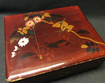 Può includere: Una scatola di legno rossa con un coperchio incernierato e una serratura. Il coperchio è decorato con un motivo floreale e di uccelli dipinto in oro, nero e bianco.