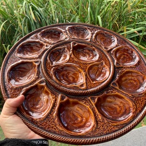 May include: A brown ceramic oyster serving platter with multiple individual shell-shaped compartments. The platter has a textured rim and a central section with additional compartments. The overall design is a rich, glossy brown.