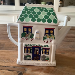 Może przedstawiać: Biały ceramiczny czajnik w kształcie domu z zielonym dachem i napisem 'tea & chip shop teapot'. Czajnik ma okna z kwiatami i drzwi z osobą w środku.
