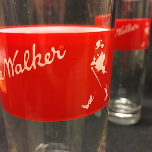 May include: Two clear glass tumblers with a red band around the middle. The band has the words "Johnnie Walker" in white script and a white silhouette of a man walking with a cane.