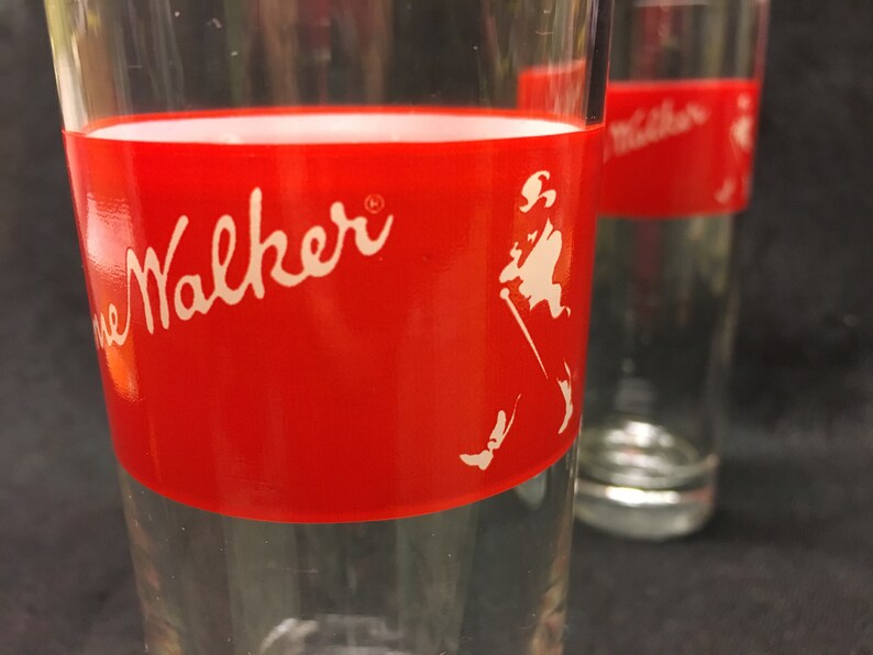 May include: Two clear glass tumblers with a red band around the middle. The band has the words "Johnnie Walker" in white script and a white silhouette of a man walking with a cane.