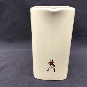 Pode incluir: Jarro de cer&acirc;mica creme com formato retangular. O jarro apresenta um pequeno bico e uma imagem decorativa de um homem com casaco vermelho e botas pretas. O jarro tem aproximadamente 18 cm de altura.