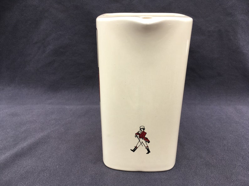 Pode incluir: Jarro de cer&acirc;mica creme com formato retangular. O jarro apresenta um pequeno bico e uma imagem decorativa de um homem com casaco vermelho e botas pretas. O jarro tem aproximadamente 18 cm de altura.