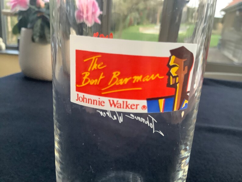 May include: A clear glass with a red, white, and blue sticker that says "The Boat Barman Johnnie Walker&reg;" with a cartoon illustration of a man's head.