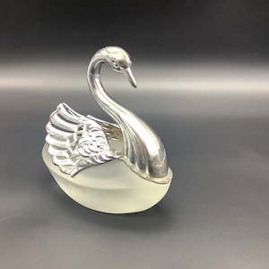 Peut inclure: Un plat décoratif en forme de cygne argenté avec une base en verre dépoli. Le cygne présente des motifs détaillés d'ailes et de plumes. Le plat est probablement utilisé pour servir ou exposer.