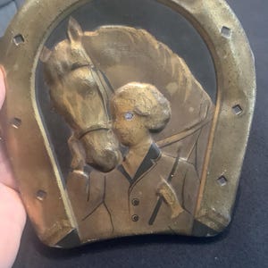 Puede incluir: Una herradura de metal de color dorado con una imagen en relieve de un caballo y una cabeza de persona en su interior. El caballo es marrón y la persona lleva una chaqueta marrón. La herradura es una decoración de pared.