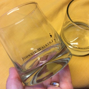 May include: Two clear glass tumblers with the Johnnie Walker logo printed on the bottom. The glasses are empty and sitting on a yellow surface.