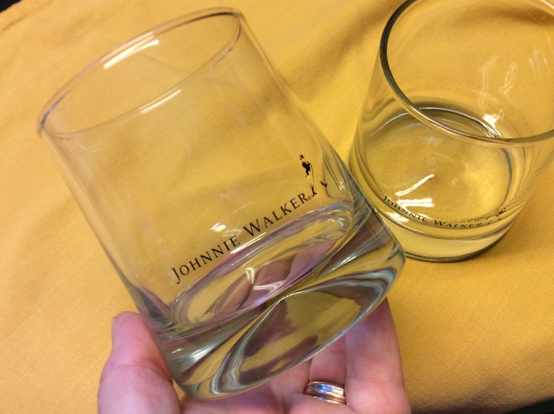 May include: Two clear glass tumblers with the Johnnie Walker logo printed on the bottom. The glasses are empty and sitting on a yellow surface.