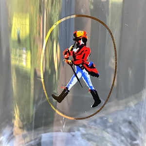 Pu&ograve; includere: Bicchiere trasparente con un design circolare bordato d'oro raffigurante una figura che cammina con un cappotto rosso, stivali neri e un cappello giallo. La figura tiene un bastone. Illustrazione classica.