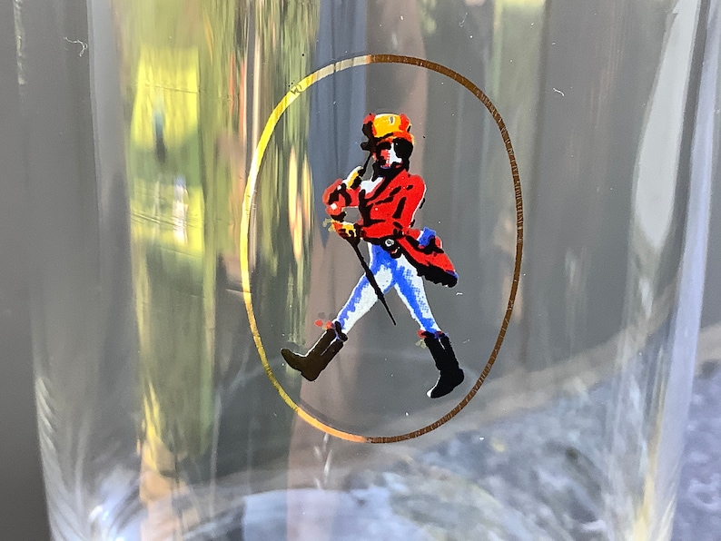 Pu&ograve; includere: Bicchiere trasparente con un design circolare bordato d'oro raffigurante una figura che cammina con un cappotto rosso, stivali neri e un cappello giallo. La figura tiene un bastone. Illustrazione classica.
