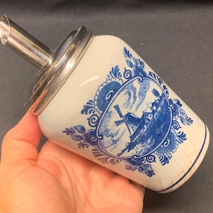 Op de afbeelding: Een wit en blauw keramisch melkkan met een windmolenontwerp. De melkkan heeft een zilveren tuit en rand. Het ontwerp toont een windmolenscène in een decoratieve blauwe rand. Een klassiek stuk voor het serveren van room of melk.