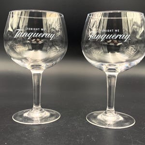 May include: Two clear, stemmed gin glasses with a rounded bowl. The text "TONIGHT WE Tanqueray" is printed in white on the front of each glass. The glasses are displayed on a dark surface.