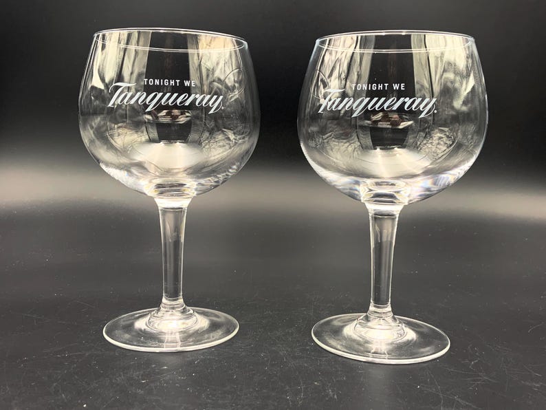 May include: Two clear, stemmed gin glasses with a rounded bowl. The text "TONIGHT WE Tanqueray" is printed in white on the front of each glass. The glasses are displayed on a dark surface.