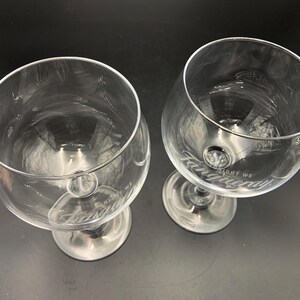 May include: Two clear, stemmed gin glasses with the word "Tanqueray" etched on the bowl. The glasses have a wide bowl and a short stem. The text "LIGHT WE" is also etched on the bowl. The glasses are empty.