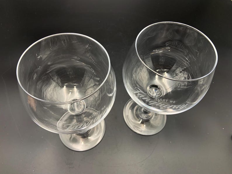May include: Two clear, stemmed gin glasses with the word "Tanqueray" etched on the bowl. The glasses have a wide bowl and a short stem. The text "LIGHT WE" is also etched on the bowl. The glasses are empty.