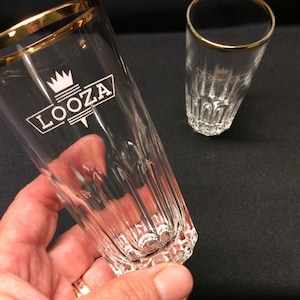 Puede incluir: Dos vasos de vidrio transparente con bordes dorados. El vaso más grande tiene la palabra "LOOZA" en texto blanco con un logotipo de corona. Los vasos tienen detalles verticales acanalados y una base facetada. Una mano sostiene un vaso.