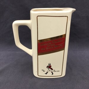 Pode incluir: Jarro retangular creme com uma al&ccedil;a, apresentando um r&oacute;tulo vermelho e dourado que diz "Johnnie Walker Red Label Old Scotch Whisky". Uma pequena ilustra&ccedil;&atilde;o de um homem andando est&aacute; abaixo do r&oacute;tulo.