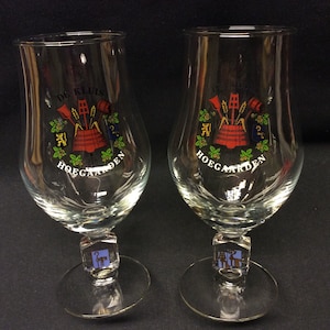 Op de afbeelding: Twee heldere Hoegaarden bierglazen. Elk glas heeft een kleurrijk embleem met tekst, waaronder "DE KLUIS" en "HOEGAARDEN". De glazen hebben een steel en een ronde voet. Het embleem bevat een rood vat en tarwe.