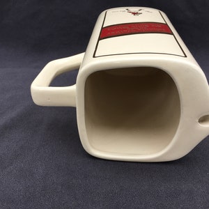 Pode incluir: Jarro de cer&acirc;mica creme com corpo retangular e al&ccedil;a curva. O jarro tem um r&oacute;tulo vermelho e preto com texto. O jarro tem um bico com um pequeno orif&iacute;cio. O jarro est&aacute; sobre uma superf&iacute;cie azul escuro.