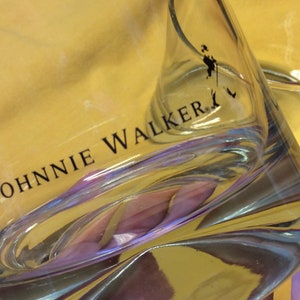 May include: A clear glass tumbler with the words "JOHNNIE WALKER" printed on the side. The glass has a textured surface and is partially filled with a yellow liquid.