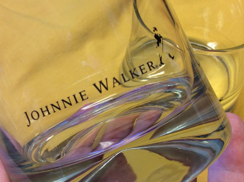 May include: A clear glass tumbler with the words "JOHNNIE WALKER" printed on the side. The glass has a textured surface and is partially filled with a yellow liquid.