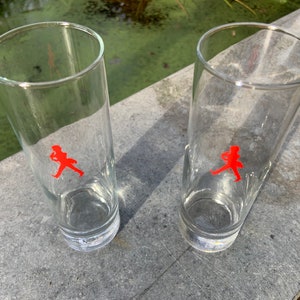 Op de afbeelding: Twee heldere glazen tumblers met een rood lopend figuurlogo op elk. De glazen staan op een grijs oppervlak.