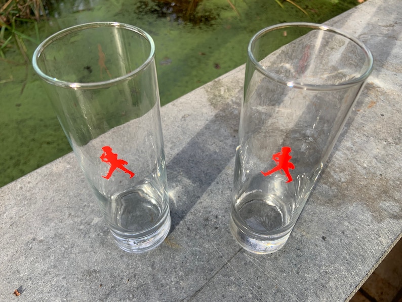Op de afbeelding: Twee heldere glazen tumblers met een rood lopend figuurlogo op elk. De glazen staan op een grijs oppervlak.