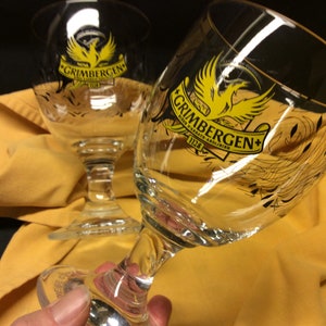 May include: Two clear Grimbergen beer glasses with a yellow and black logo. The logo features a bird and the text "Grimbergen" and "1128". The glasses are on a yellow cloth.