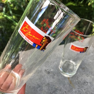 May include: Two clear glass drinking glasses with red and blue labels that say "The  But Barman Johnnie Walker" and feature a cartoon illustration of a man with a mustache.